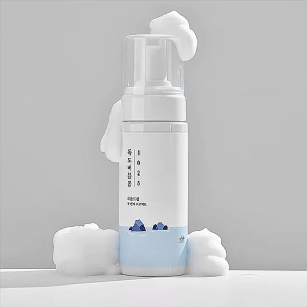 Un flacon blanc de mousse nettoyante Dokdo Round Lab 1025 de 150 ml, muni d'une pompe, libère une mousse blanche et onctueuse. Des motifs de montagnes bleues et un texte coréen ornent le flacon, entouré de mousse sur une surface gris clair. Dakar