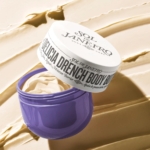 Un pot violet ouvert de Sol de Janeiro Delicia Drench 75 ml révèle un beurre corporel crémeux à l'intérieur, reposant sur un fond beige clair texturé de la crème riche, rappelant l'emblématique crème brésilienne Bum Bum. Dakar
