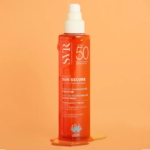 Un flacon de Svr Sun Secure Huile SPF50 Protection Solaire Format Huile non Grasse 200 ml se dresse debout sur une surface orange avec de l'huile renversée à sa base, sur un fond orange vif. Dakar