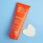 Un tube orange de SVR Sun Secure Lait Solaire Hydratant SPF50+ Fini Invisible 100 ml repose sur une surface bleue à côté de deux bandes de lotion blanches, mettant en valeur la haute protection offerte par cet écran solaire hydratant. Dakar