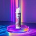 Une boîte étiquetée « Tickle My Body » est posée sur un plateau circulaire, entourée de reflets lumineux irisés et colorés sur fond violet. L'emballage est rose et blanc, avec une pompe doseuse. Dakar