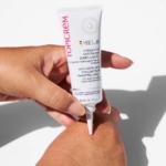 Une main presse un tube de Topicrem Mela Soin Corps Concentré Anti Taches Zones Ciblées 50 ml, déposant la crème sur le dos d'une autre main sur fond blanc. Dakar