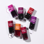 Huit bouteilles d'Etude Encre à Lèvres Effet Naturel Dear Darling Water Tint dans des tons rouges, orange et violets sont présentées sur une surface blanche avec des gouttelettes d'eau claires dispersées autour. Dakar