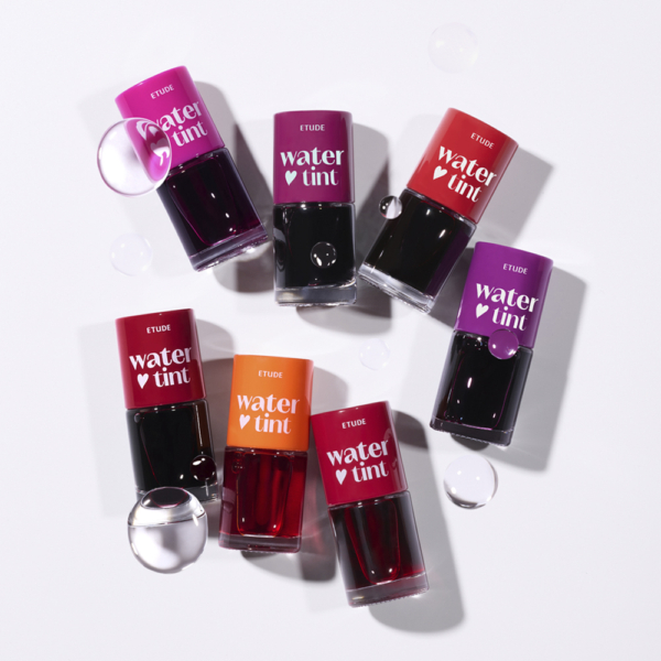 Huit bouteilles d'Etude Encre à Lèvres Effet Naturel Dear Darling Water Tint dans des tons rouges, orange et violets sont présentées sur une surface blanche avec des gouttelettes d'eau claires dispersées autour. Dakar