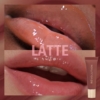 Gros plan sur des lèvres portant un brillant nude-marron Makeup Revolution Juicy Peptide Baume à Lèvres avec des Peptides 8 ml, avec la mention "LATTE" en surimpression. Le tube de gloss apparaît dans le coin inférieur droit. Dakar