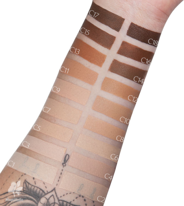 Un bras présente 16 échantillons rectangulaires des teintes de Makeup Revolution Conceal and Define Correcteur Liquide Visage et Contour Yeux 4 g, étiquetés de C1 à C18, de très clair à foncé, avec un tatouage floral visible près du poignet. Dakar
