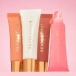Quatre tubes de Makeup Revolution Juicy Peptide Baume à Lèvres avec des Peptides 8 ml avec des bouchons dorés sont présentés sur un fond rose - trois debout en pêche, blanc et mauve, et un ouvert en rose avec du produit pressé. Dakar