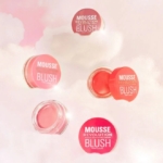 Cinq pots de Makeup Revolution Mousse Blush Fard à Joues 6 g dans des teintes roses et corail sont présentés sur un fond rose doux, semblable à un nuage, et plusieurs d'entre eux sont ouverts pour révéler le blush crémeux à l'intérieur. Dakar