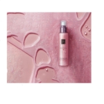 Un flacon rose RITUALS The Ritual of Sakura Brume Scintillante pour Corps 150ml repose sur une surface rose étincelante avec des gouttelettes d'eau, créant une scène coordonnée et chatoyante. Dakar
