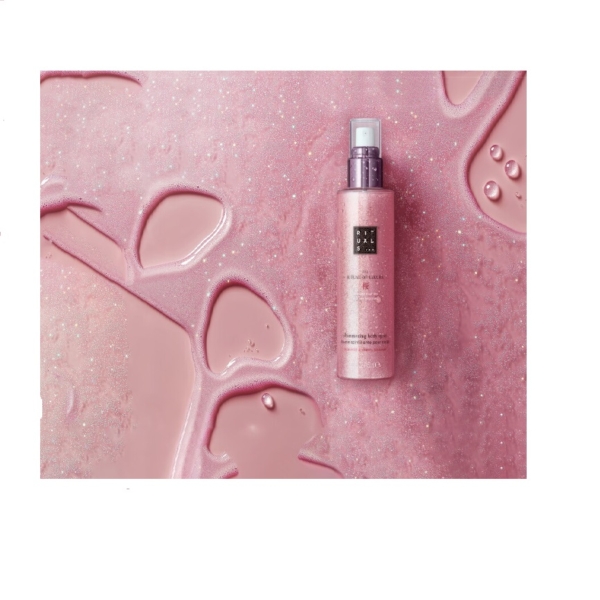 Un flacon rose RITUALS The Ritual of Sakura Brume Scintillante pour Corps 150ml repose sur une surface rose étincelante avec des gouttelettes d'eau, créant une scène coordonnée et chatoyante. Dakar