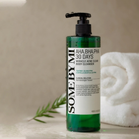 Une bouteille verte de Some By Mi Mousse Nettoyante Pour le Corps Anti Acné 30 Jours Miracle 400 g repose sur une surface beige à côté d'une serviette blanche roulée et d'une petite tige de plante verte. Dakar