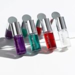 Six flacons de Tirtir Huile à Lèvres Effet Naturel My Glow Lip Oil 5,7 ml dans des tons violets, bleu sarcelle, rouges, verts, orange et clairs vibrants sont disposés sur une surface blanche avec des ombres et des reflets doux. Dakar