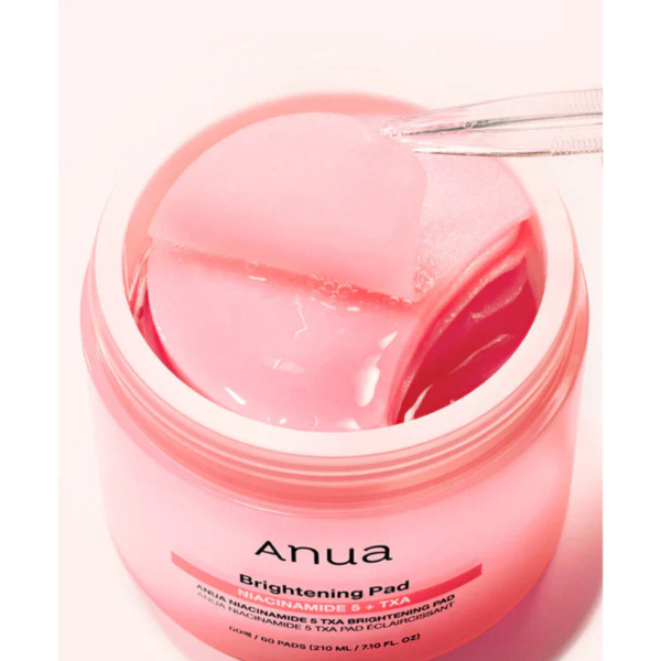 Gros plan sur le pot rose d'Anua Pads Anti Taches et Imperfections à la Niacinamide 5 TXA Brightening, rempli de coussinets roses ronds. Une pince à épiler permet de soulever un coussinet, et l'étiquette indique que la niacinamide et le TXA font partie des ingrédients clés. Dakar
