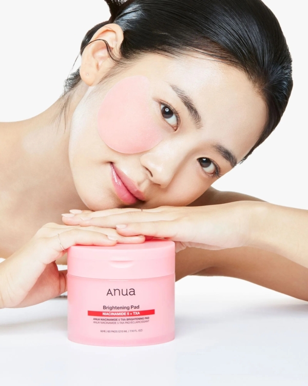 Une femme à la peau claire sourit doucement en posant son menton sur ses mains. Un tampon de soin rose est posé sur sa joue et un pot rose étiqueté "Anua Pads Anti Taches et Imperfections à la Niacinamide 5 TXA Brightening 60 pads" est posé devant elle sur un fond blanc. Dakar