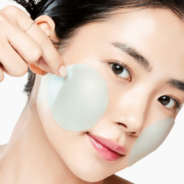 Une femme à la peau éclatante applique délicatement Anua PDRN Hydratant Éclat Visage Anti Age 100 Hyaluronic Acid Glow 60 Pads sur ses joues pendant sa routine de soins, en tenant un patch d'hydrogel bleu clair et en regardant la caméra. Dakar