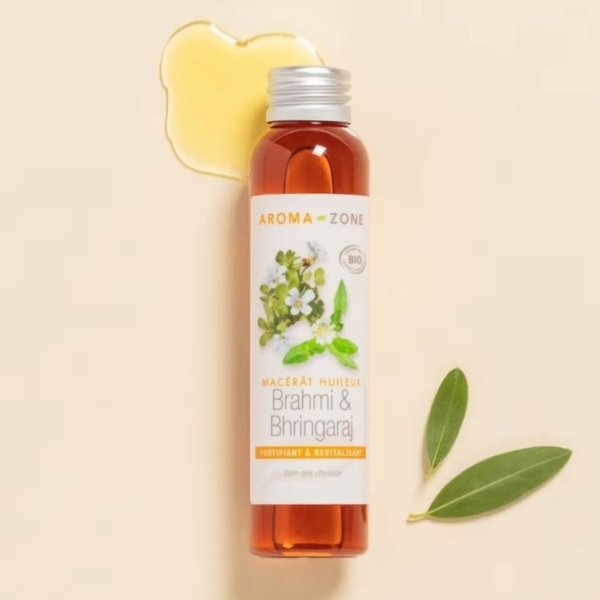 Un flacon d'Aroma Zone Huile de Brahmi et Bhringaraj Bio (Macérât Huileux) 250 ml repose sur une surface légère avec une petite goutte d'huile au-dessus et deux feuilles vertes à côté. L'étiquette présente des illustrations botaniques des ingrédients. Dakar