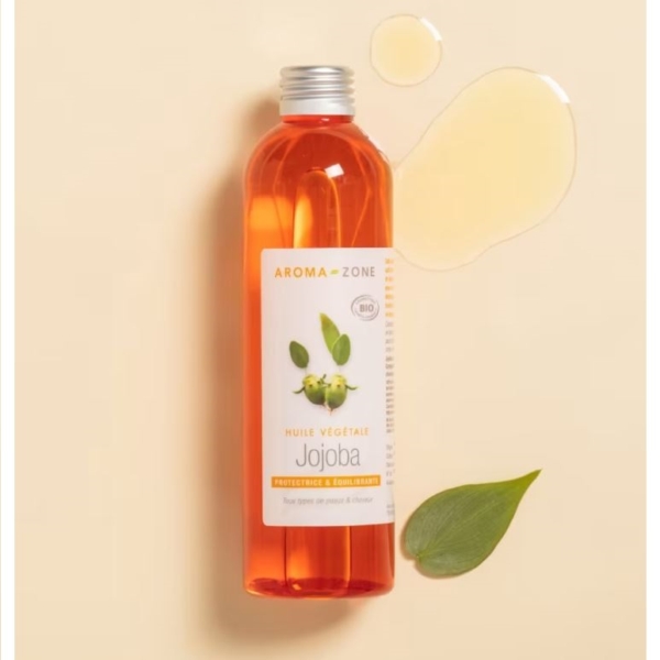 Un flacon d'Aroma Zone Huile de Jojoba BIO 250 ml avec un bouchon orange est présenté sur un fond beige à côté d'une feuille verte et d'une petite flaque d'huile. Dakar