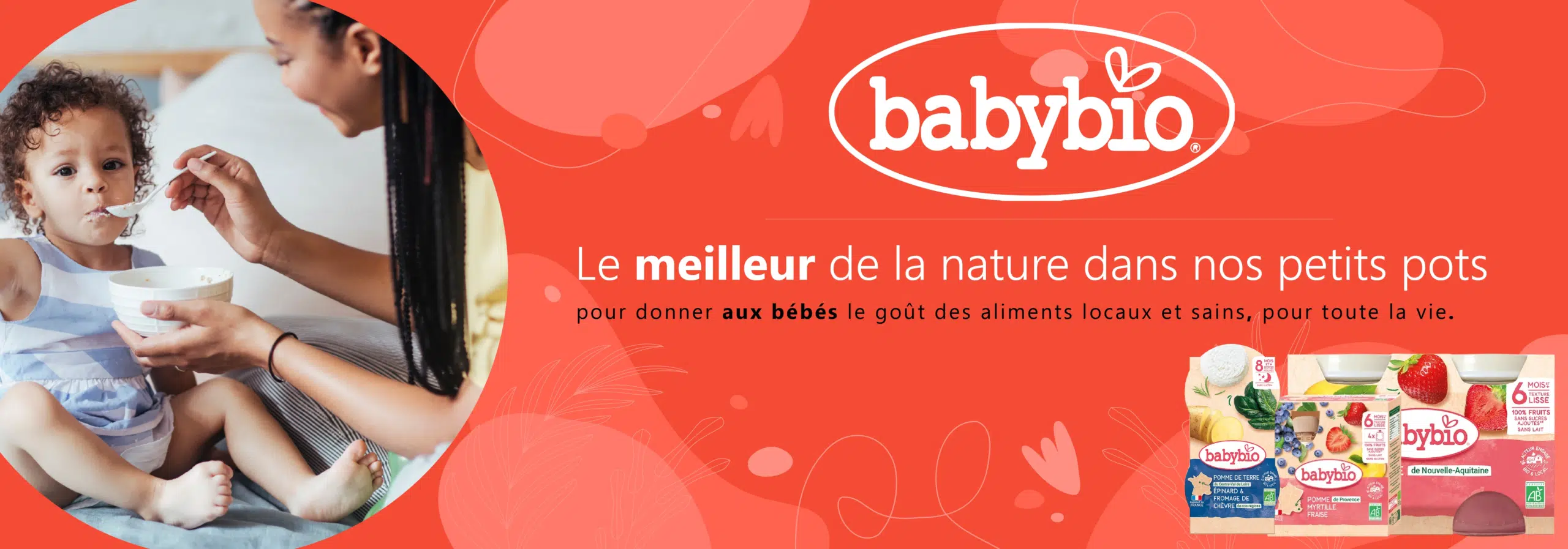 Une mère nourrit son jeune enfant à la cuillère. À côté d'eux, une publicité de Babybio et Univers Cosmetix, qui fait la promotion des aliments biologiques pour bébés, avec le slogan "Le meilleur de la nature dans nos petits pots" et des images des produits Babybio. Dakar