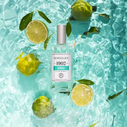 Une bouteille de Berdoues 1902 Neroli Eau de Cologne par Univers Cosmetix repose dans une eau bleue claire où flottent des moitiés de citron vert, des agrumes verts et des feuilles. La lumière du soleil crée des ondulations et des reflets à la surface de l'eau. Dakar