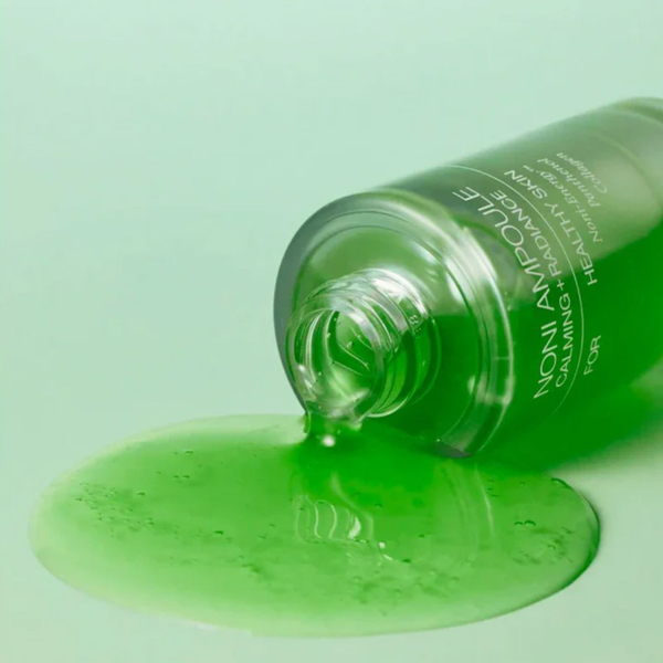 Un flacon de 30 ml de Celimax Ampoule Éclat Anti Age The Real Noni Energy étiqueté "NONI AMPOULE" est couché sur le côté, le liquide vert vibrant se déversant sur une surface assortie, soulignant les effets apaisants, l'éclat et la santé de la peau. Dakar