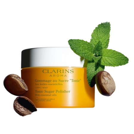 Un pot d'Aroma Gommage Corps au Sucre Tonic aux Huiles Essentielles 250 ml de Clarins est présenté avec deux noix de karité et un brin de menthe fraîche sur fond blanc. Dakar