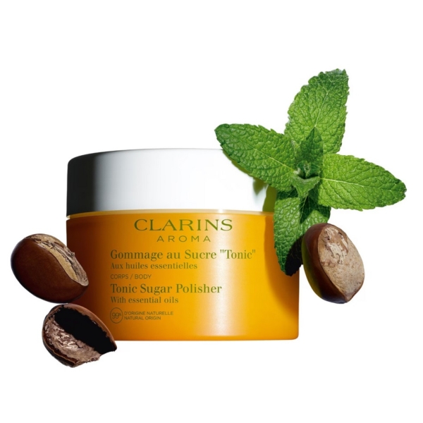 Un pot d'Aroma Gommage Corps au Sucre Tonic aux Huiles Essentielles 250 ml de Clarins est présenté avec deux noix de karité et un brin de menthe fraîche sur fond blanc. Dakar