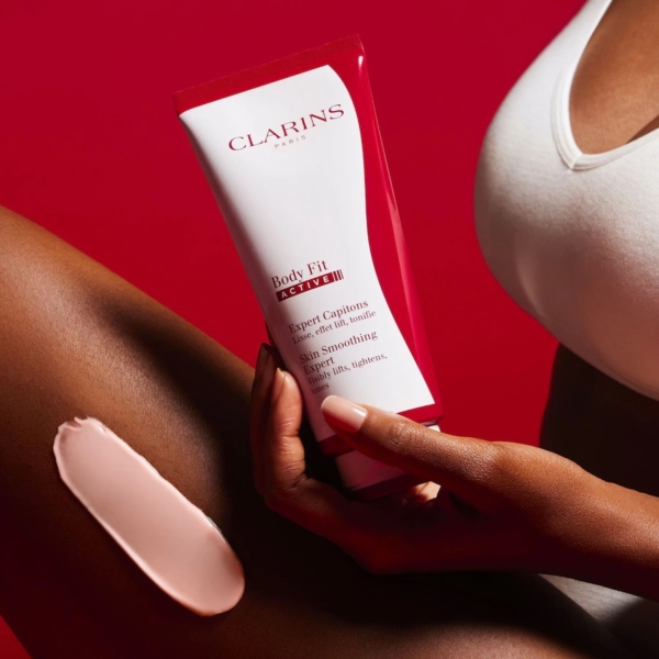 Une personne vêtue d'un débardeur blanc applique la Crème Corps Tonifiant Active Fit Anti Age Peau Lisse 200 ml de Clarins sur sa cuisse, en tenant le tube rouge et blanc sur un fond rouge, avec une trace de crème visible sur sa peau. Dakar