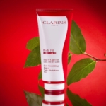 Un tube rouge et blanc de Crème Corps Tonifiant Active Fit Anti Age Peau Lisse 200 ml de Clarins se tient debout sur une surface brillante avec des feuilles vertes en arrière-plan, sur une toile de fond rouge vif. Dakar
