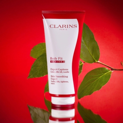 Un tube rouge et blanc de Crème Corps Tonifiant Active Fit Anti Age Peau Lisse 200 ml de Clarins se tient debout sur une surface brillante avec des feuilles vertes en arrière-plan, sur une toile de fond rouge vif. Dakar