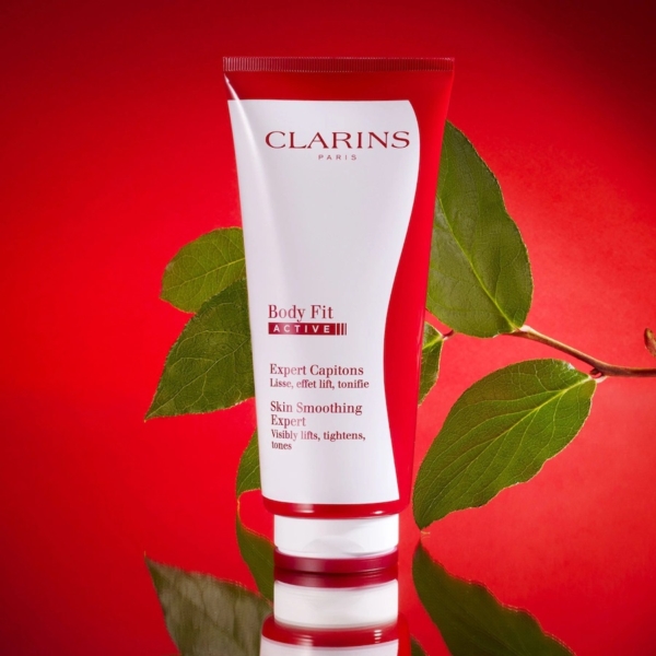Un tube rouge et blanc de Crème Corps Tonifiant Active Fit Anti Age Peau Lisse 200 ml de Clarins se tient debout sur une surface brillante avec des feuilles vertes en arrière-plan, sur une toile de fond rouge vif. Dakar