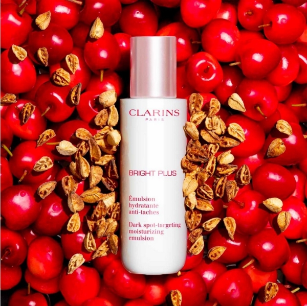 Un flacon d'Emulsion Hydratante Anti Taches Bright Plus 75 ml de Clarins est posé sur un lit de baies rouge vif et de graines éparses, créant un arrière-plan vibrant et accrocheur. Dakar