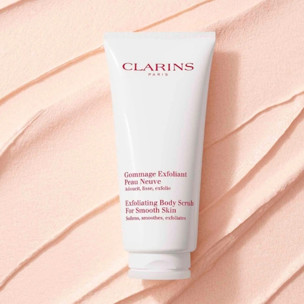 Un tube blanc de Clarins Gommage Corps Peau Neuve et Lisse 200 ml avec un texte rouge et or est placé sur un fond texturé rose pâle avec des lignes ondulées. Dakar