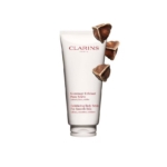 Un tube de Gommage Corps Peau Neuve et Lisse 200 ml de Clarins est posé sur un fond blanc, avec à ses côtés des morceaux d'exfoliant marron. Dakar