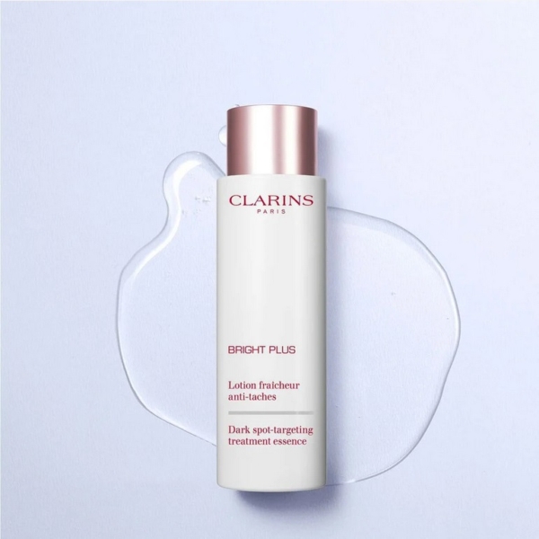 Un flacon de l'Essence de Traitement Anti-Taches Clarins Bright Plus se tient debout sur une surface claire, avec un liquide translucide en dessous. Le flacon a un corps blanc et un bouchon rose métallique. Dakar