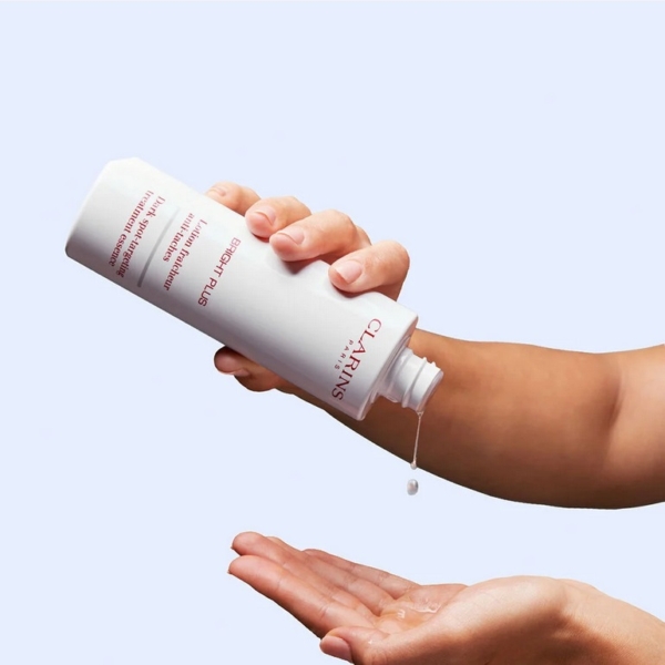 Une main verse le liquide d'un flacon Clarins blanc étiqueté "Bright Plus" sur la paume d'une autre main, sur un fond bleu clair. Dakar