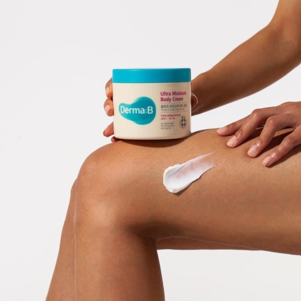 Une personne applique Derma B Crème Corps Ultra Hydratante et Apaisante Peau Sèche 430 ml sur sa jambe, tenant son pot avec un couvercle et une étiquette bleus sur un fond blanc uni. Dakar