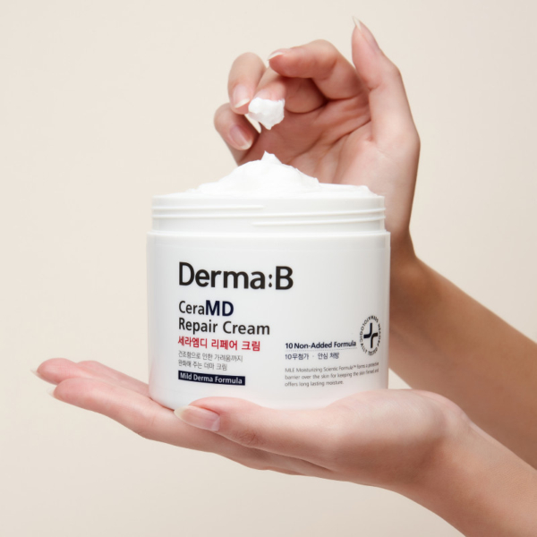 Une main tient un flacon ouvert de Derma B Crème Réparatrice et Nourrissante pour le corps 430 ml tandis qu'une autre prélève la crème dans le pot, sur un fond beige uni. Dakar