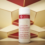 Un flacon de 1 litre de HT26 Gel Douche Gommant Multi Éclaircissant se tient debout sur un fond géométrique rouge et or. Le flacon blanc est orné d'un texte rouge et or, soulignant ses bienfaits exfoliants et éclaircissants pour la peau. Dakar