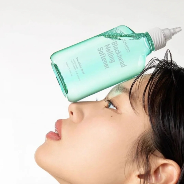 Une femme aux cheveux mouillés est allongée sur le dos, tenant en équilibre sur son nez un grand flacon de Jumiso Kit Soins avec 1 Adoucissant Anti Points noirs 150 ml et 40 Cotons, tandis que le liquide se déverse sur sa peau, sur un fond blanc. Dakar