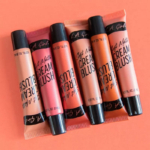 Cinq tubes de L.A Girl Blush Lèvres et Joues Soft Matte Cream Blush 8 ml dans des teintes assorties de rose, rouge et pêche sont disposés en diagonale sur un fond pêche. Dakar