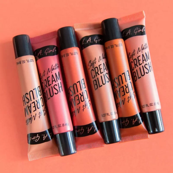 Cinq tubes de L.A Girl Blush Lèvres et Joues Soft Matte Cream Blush 8 ml dans des teintes assorties de rose, rouge et pêche sont disposés en diagonale sur un fond pêche. Dakar
