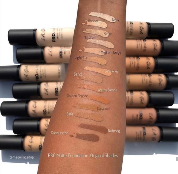 Un bras présente des échantillons de L.A Girl Fond Teint Crémeux Haute Couvrance Longue Tenue 30 ml dans des teintes allant d'Ivoire et Bisque à Muscade et Cappuccino, avec des flacons de fond de teint disposés à l'arrière-plan. Dakar