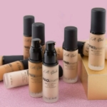 Plusieurs flacons de L.A Girl Fond Teint Crémeux Haute Couvrance Longue Tenue 30 ml dans différentes teintes sont disposés sur une surface rose, certains debout et d'autres couchés, deux étant élégamment présentés sur une boîte dorée. Dakar