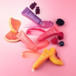 Quatre tubes de Laneige Lip Glowy Baume Brillant pour les Lèvres 10 g sont disposés sur un fond rose avec des tranches de pamplemousse, de pêche, des baies assorties, des oursons en gomme et des échantillons de baumes teintés. Dakar