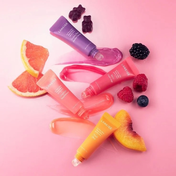 Quatre tubes de Laneige Lip Glowy Baume Brillant pour les Lèvres 10 g sont disposés sur un fond rose avec des tranches de pamplemousse, de pêche, des baies assorties, des oursons en gomme et des échantillons de baumes teintés. Dakar