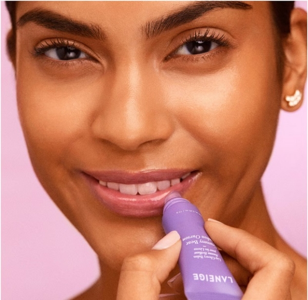 Une femme à la peau lisse sourit doucement en appliquant le Lip Glowy Baume Brillant pour les Lèvres 10 g de Laneige sur ses lèvres, portant des boucles d'oreilles en or sur un fond rose tendre. Dakar