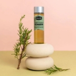 Un flacon de Macérat huileux de Romarin BIO 75ml repose sur deux pierres beiges et lisses, avec à ses côtés une branche de romarin, sur un fond rose et jaune. Dakar