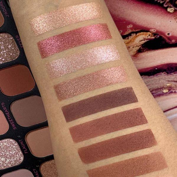 Un bras montre des échantillons de dix teintes - des roses et des ors pailletés aux bruns mats - à côté d'une Palette de Fards à Paupières Forever Flawless Allure 18 Nuances de MakeUp Revolution ouverte avec des couleurs assorties. Dakar