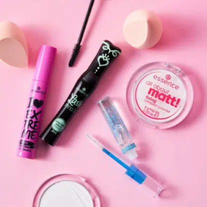 Des produits cosmétiques d'Univers Cosmetix et d'Essence, notamment du mascara, du vernis à ongles, de la poudre, du rouge à lèvres et une brosse à maquillage, sont disposés proprement sur une surface rose. L'emballage aux couleurs vives ajoute une touche d'animation à la présentation. Dakar