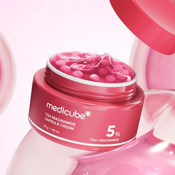 Un pot rose ouvert de Medicube Crème Capsule TXA Niacinamide 55 g révèle des capsules roses à l'intérieur. L'étiquette indique "5% TXA + Niacinamide" et elle est entourée de sphères roses douces et brillantes. Dakar